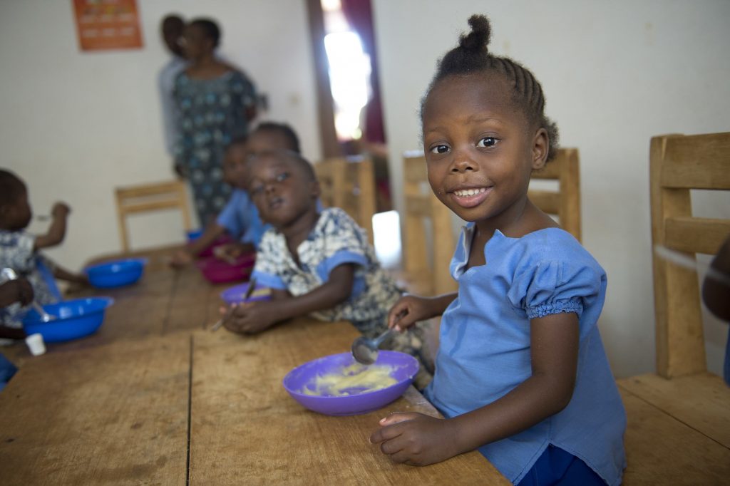 A young girl named Dieu-Merci smiles at the camera while eating her meal in the orphanage