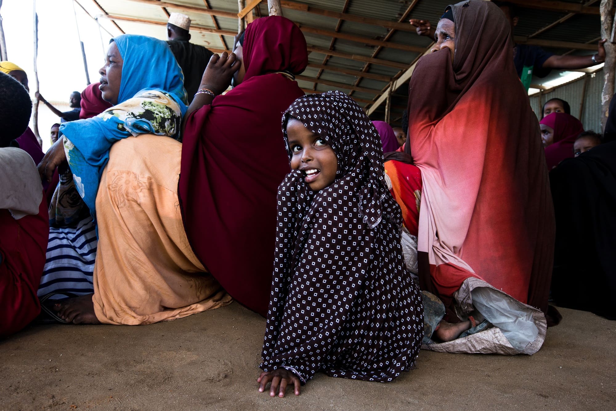 A young girl sits in a line at a food center, wearing a polka dot hijab over her head