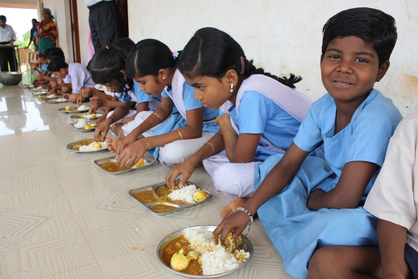 Young girls in blue school uniforms sit on the floor to eat a meal.