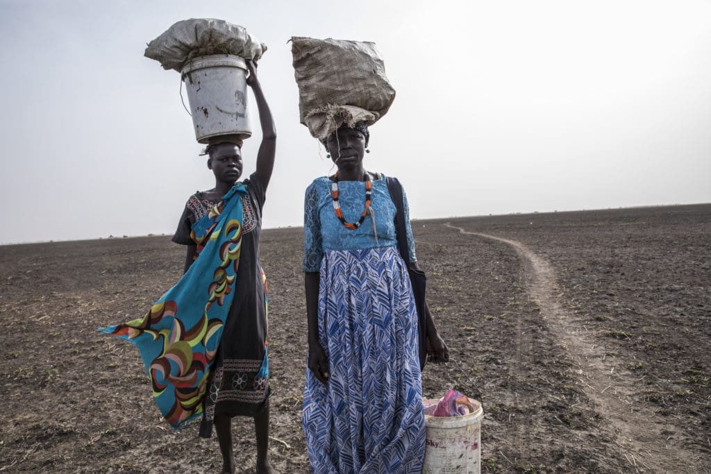 Two women carry crops across a dry field
