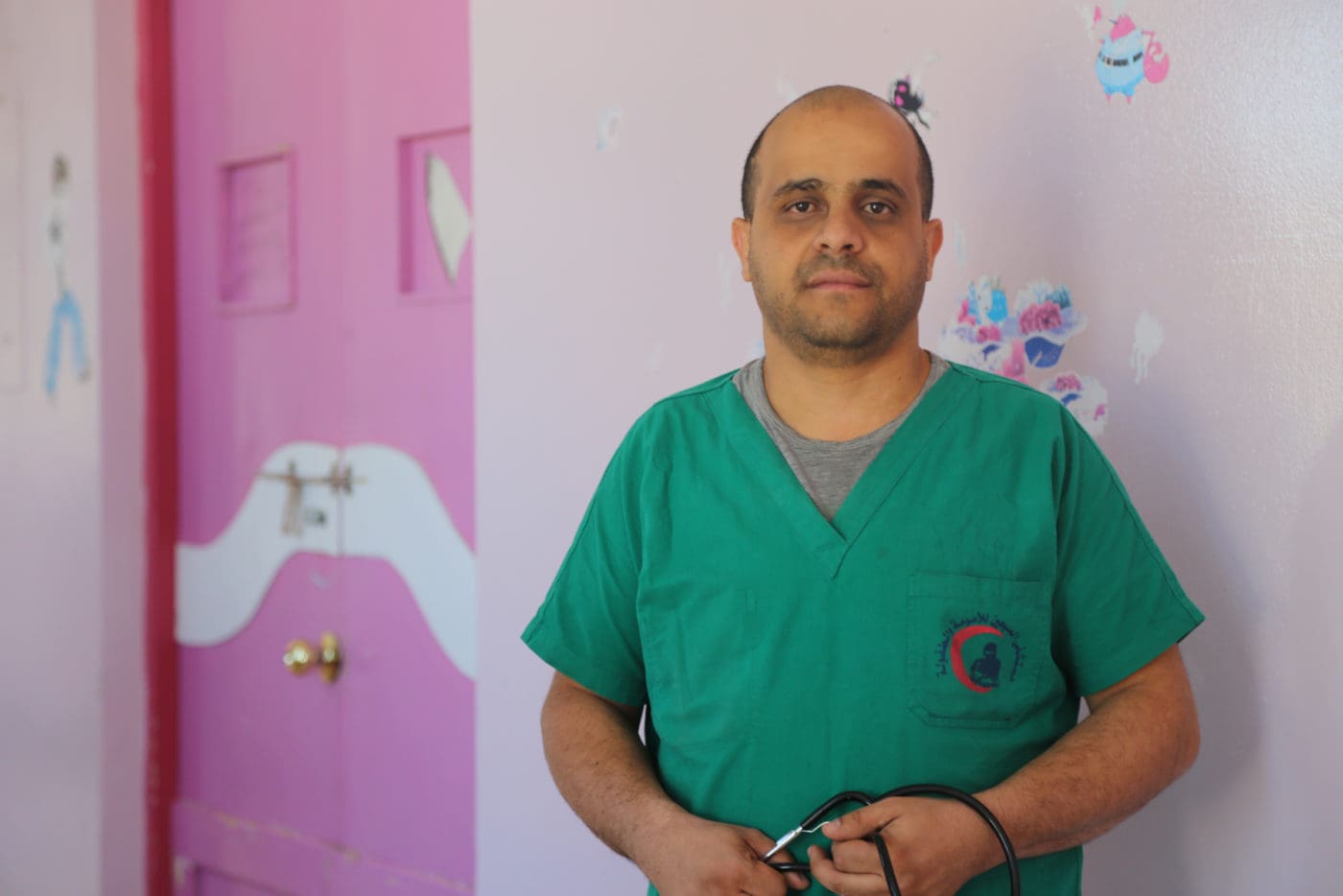 A nurse in green scrubs stands in front of a pink wall