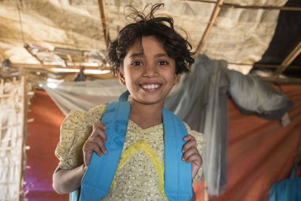 A young girl standing in a tent in a yellow backpack smiles at the camera