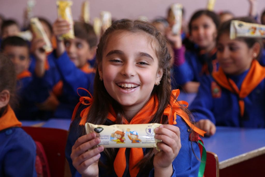 A young girl in a school uniform holds a wrapped bar up to the camera, smiling.
