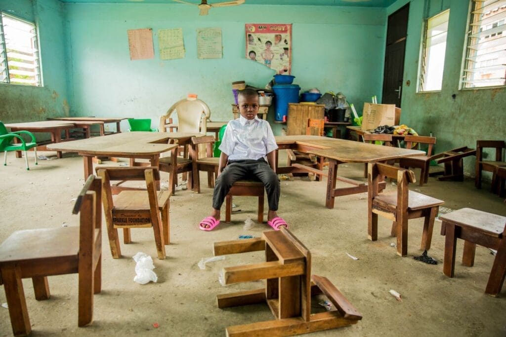 A young boy sits in a chair in an empty classroom, wearing a school uniform, looking straight at the camera.