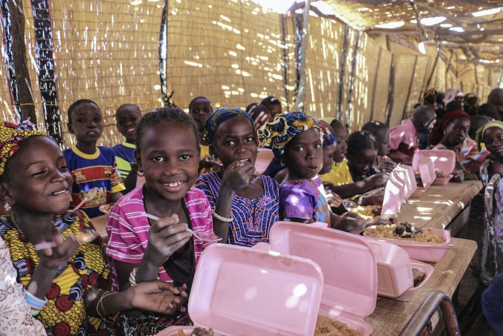 School children sit in rows at tables, holding pink boxes of food