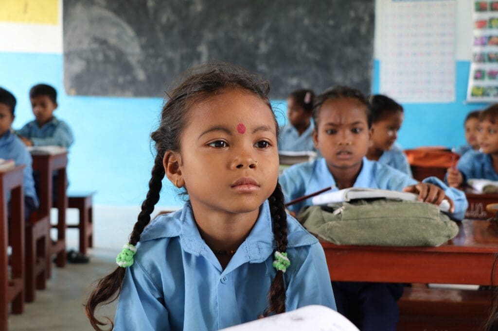 A young girl in a school uniform sits at a school desk in a classroom.