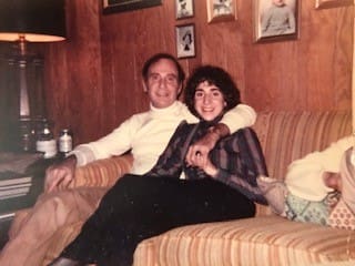 A young woman sits on a couch next to her father, his arm around her.