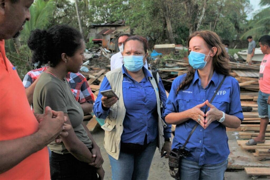 WFP workers in masks talk to a man and woman, surrounded by hurricane rubble.