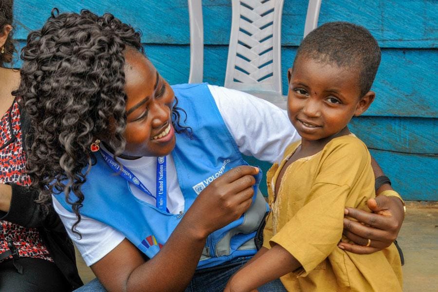 A woman in a UN volunteer vest smiles and poses for a photo with a young child.
