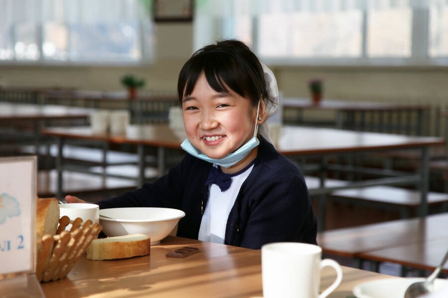 A schoolgirl sits at a table to eat a meal, smiling, mask under her chin.
