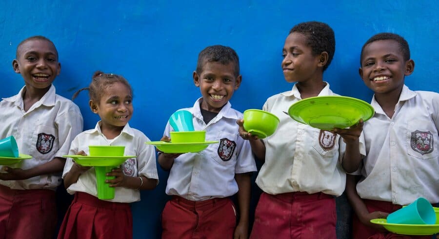 5 schoolkids pose in front of a blue wall, holding green plates and cups.