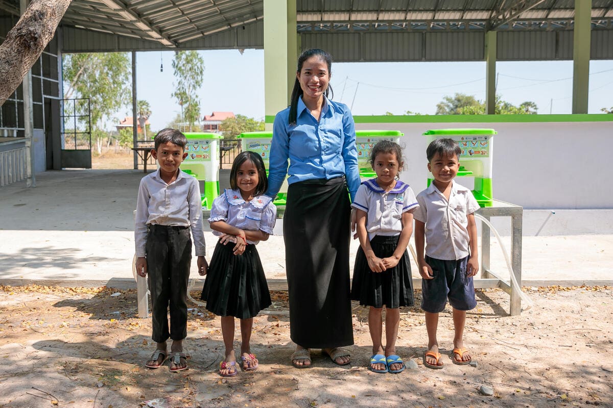 A teacher smiles, standing with four young students.
