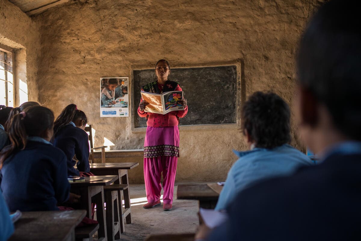 A teacher stands in front of a classroom of students, reading from a book.