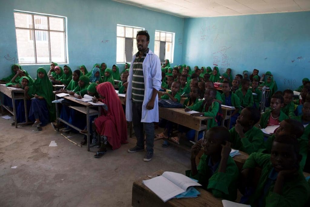 A teacher in a white coat stands in front of a classroom of students in green uniforms.