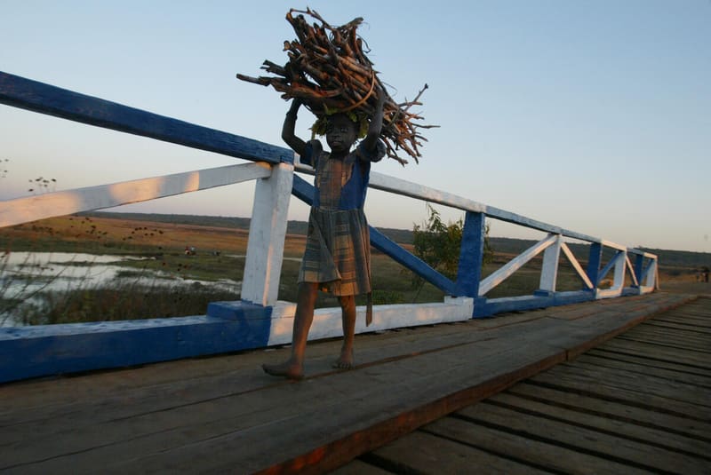 A young boy walks across a bridge, carrying a bundle of sticks on his head